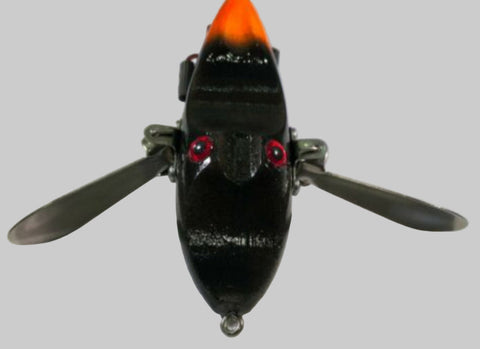 A black topwater fishing bait with wings and an orange tail pictured in a white background.